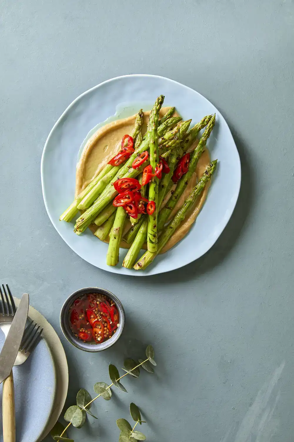Ekoplaza-biologisch-LekkerWeten-Lente2026-groeneasperges-misosaus Een gerecht met groene asperge met misosaus en rode chilipickle op een blauwe achtergrond