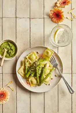 Ekoplaza-biologisch-LekkerWeten-Zomer2024-Bioverde-gevuldepasta-ravioli-saliepesto Een bord met ravioli met saliepesto op roze tegels met een vaasje met bloemen